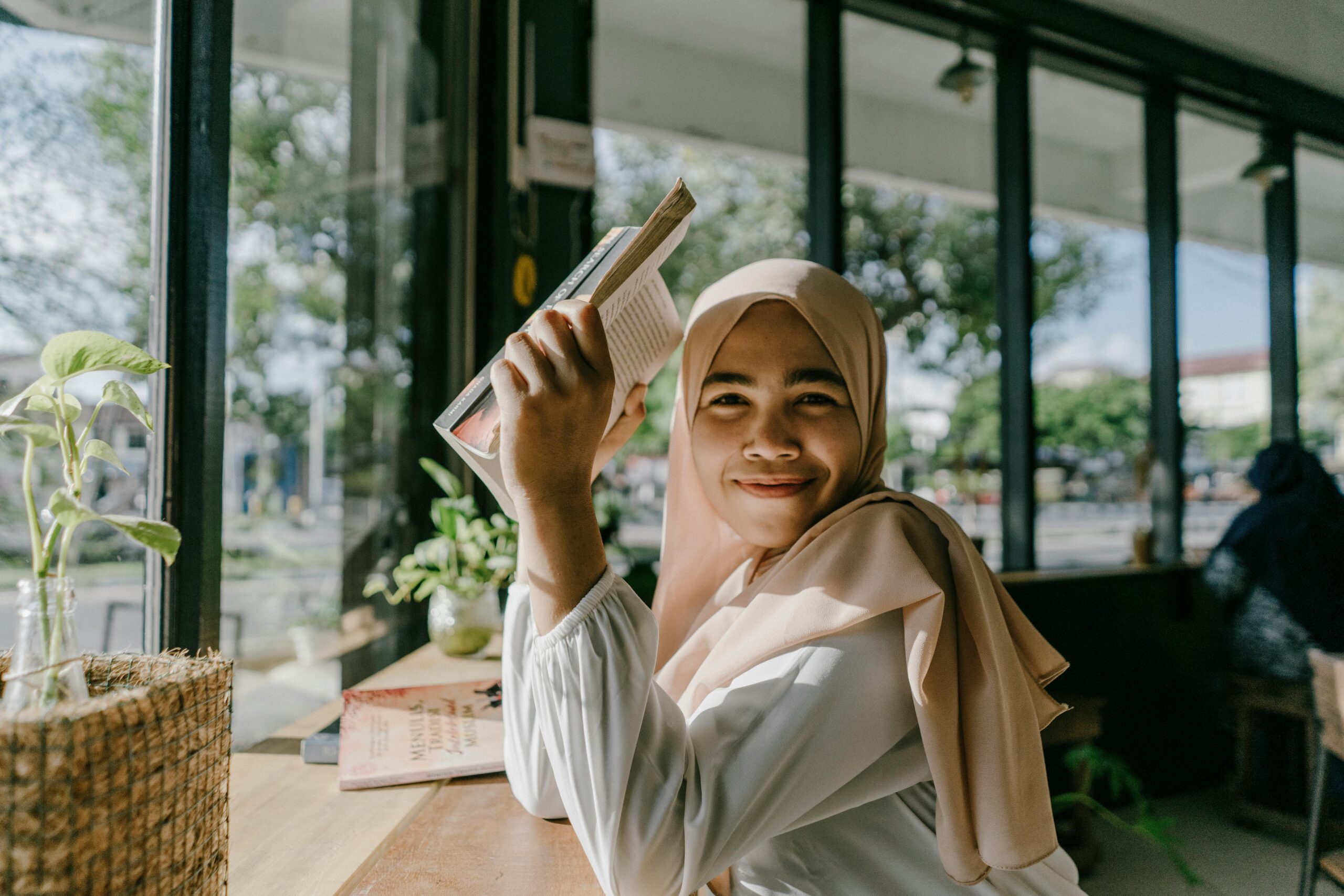 A smiling woman in a hijab reading a book by a window in a cozy cafe in Banda Aceh, Indonesia.